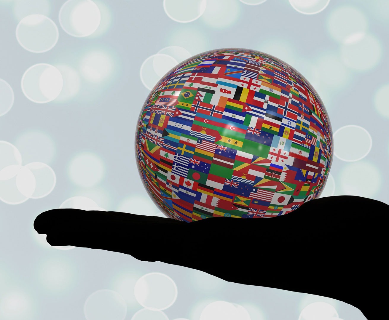 Ausgestreckte Hand im Schatten mit Ball in der Hand, welcher vollständig mit unterschiedlichen kleinen Landesflaggen bedeckt ist, als Symbol für die Vielfalt der Sprachen