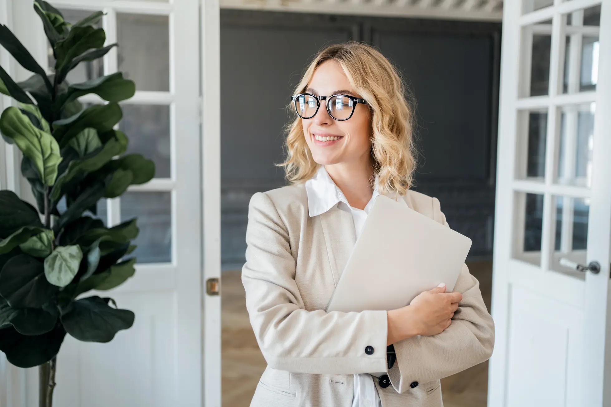Fröhliche Geschäftsfrau in einem beigen Blazer, die eine Aktentasche hält und optimistisch zur Seite blickt, in einem hellen Büro mit einer grossen Zimmerpflanze neben einer Glastür.