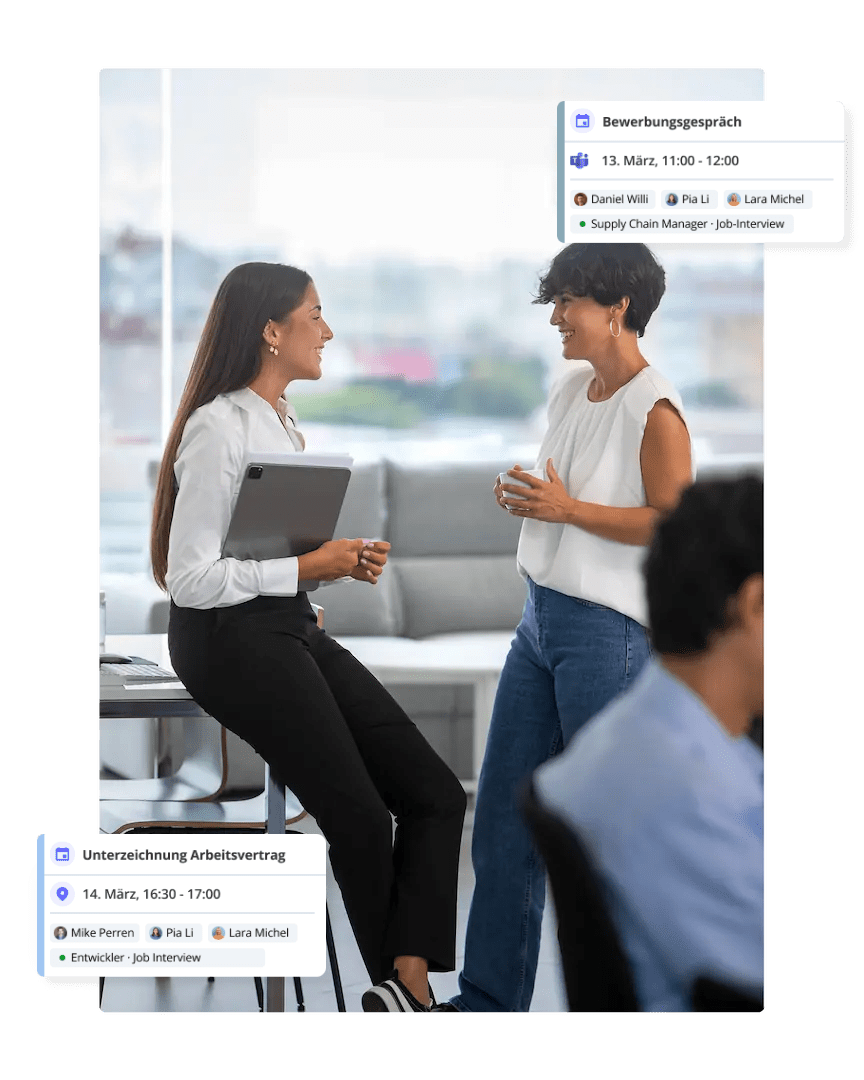 Zwei Geschäftsfrauen unterhalten sich neben einem Laptop, auf dessen Bildschirm im Hintergrund Kalenderereignisse wie Vorstellungsgespräche angezeigt werden