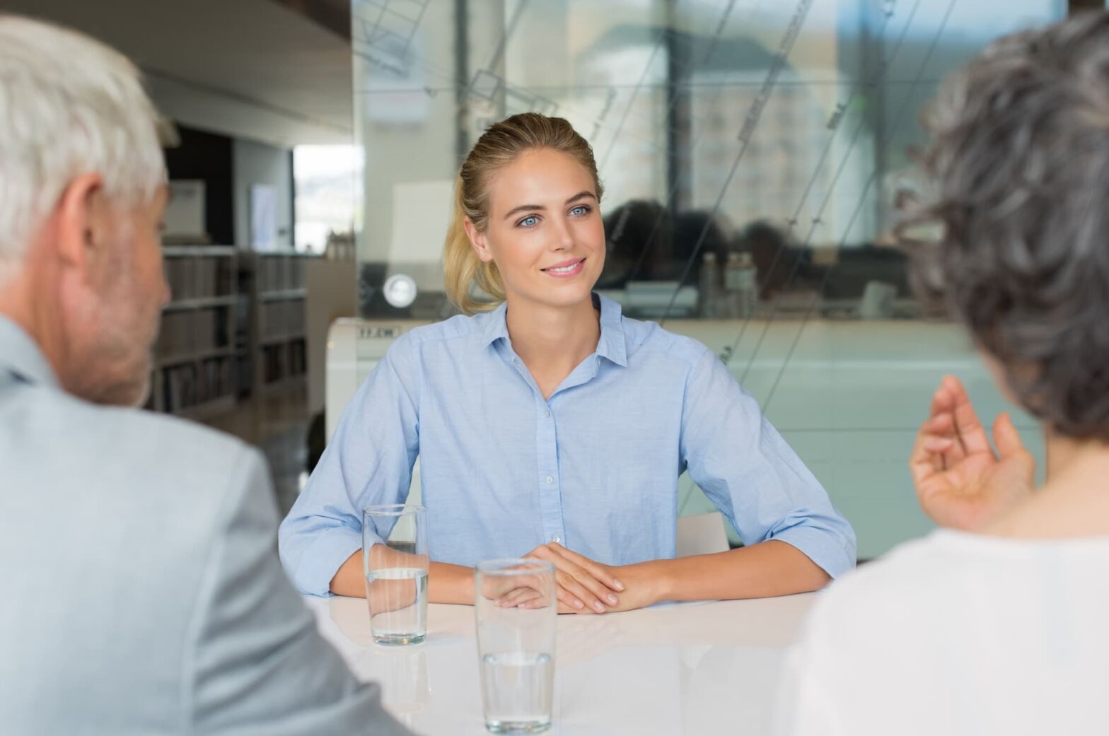 A young woman in a blue shirt sits at a table and takes part in a job interview. Sitting opposite her are two older people asking questions. There are glasses of water on the table.